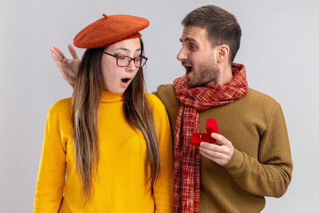 Free photo young beautiful couple happy man making proposal with engagement ring in red box to his amazed girlfriend in beret during valentines day standing over white wall