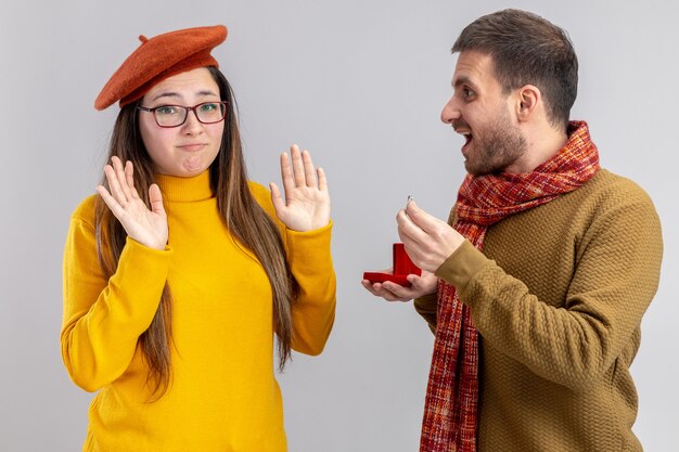 young beautiful couple happy man making proposal with engagement ring in red box to his confused and displeased girlfriend in beret during valentines day standing over white wall