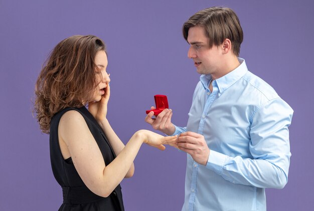 young beautiful couple happy man making a proposal with engagement ring in red box for his lovely surprised girlfriend during valentines day standing over blue background