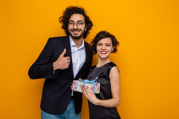 Young beautiful couple happy man showing thumbs up and woman with present looking at camera smiling cheerfully celebrating international women's day march 8 standing over orange background