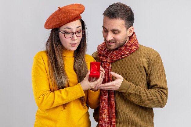 若い美しいカップルの幸せな男と白い背景の上に立っているバレンタインデーを祝う赤いボックスの婚約指輪とベレットの笑顔の女性