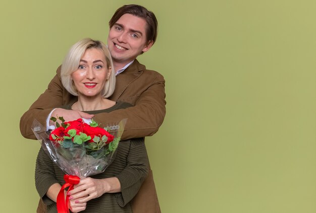 Молодая красивая пара счастливый мужчина с букетом красных роз и женщина, обнимающая счастливая в любви, вместе празднует международный женский день, стоя над зеленой стеной