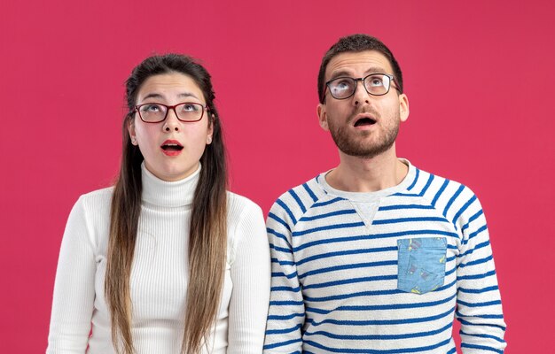 young beautiful couple happy man and woman in casual clothes wearing glasses looking up happy and surprised celebrating valentines day concept standing over pink wall