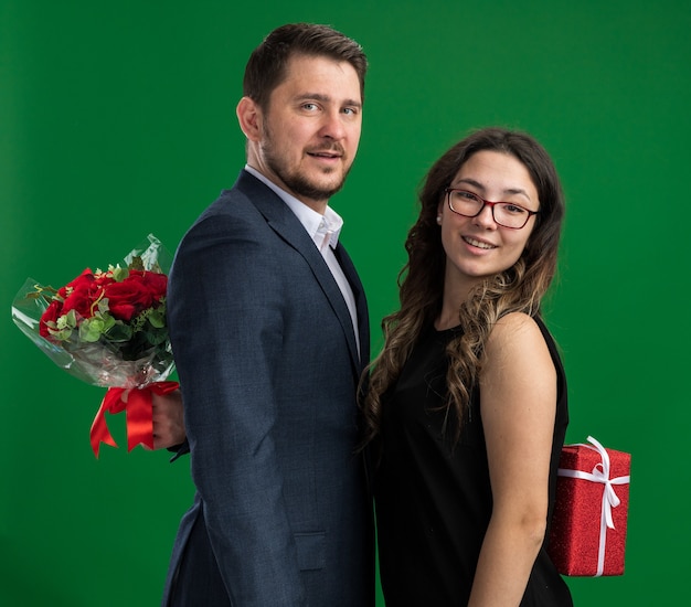 Young beautiful couple happy man and woman hiding gifts from each other happy in love together celebrating valentines day standing over green wall