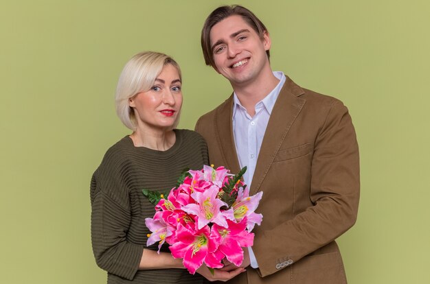 緑の壁の上に立って国際女性の日を祝って一緒に笑顔で幸せを抱きしめる花の花束を持つ若い美しいカップル幸せな男と女