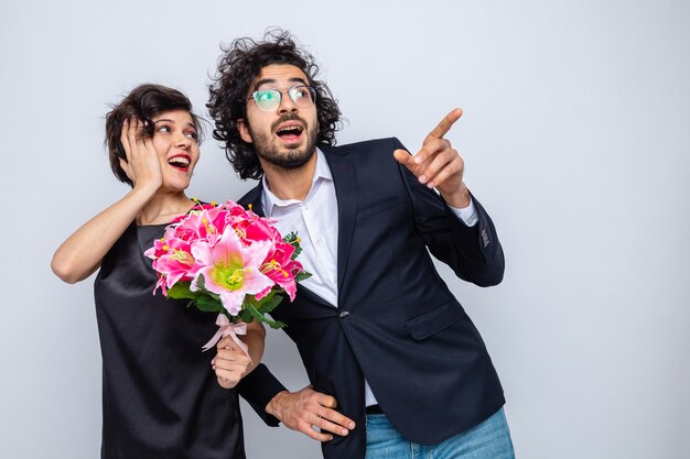Молодая красивая пара, счастливый мужчина и женщина с букетом цветов, глядя в сторону, счастливые и удивленные, указывая указательными пальцами в сторону, празднуя международный женский день 8 марта