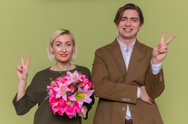 Young beautiful couple happy man and woman with bouquet of flowers looking at front smiling cheerfully showing v-sign celebrating international women's day standing over green wall