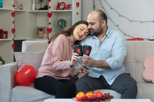 Young beautiful couple happy man and woman with coffee cups happy in love together embracing celebrating international women's day sitting on a couch in light living room