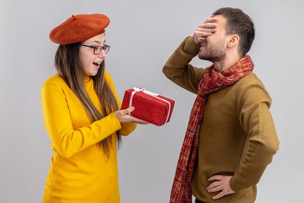 Foto gratuita giovane bella coppia donna felice in berretto dando un regalo per il suo fidanzato che copre gli occhi con le mani felici innamorati insieme celebrando il giorno di san valentino in piedi sul muro bianco