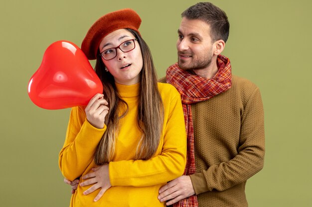 молодая красивая пара счастливая женщина в берете с воздушным шаром в форме сердца и счастливый мужчина с шарфом празднует день святого валентина, стоя на зеленом фоне