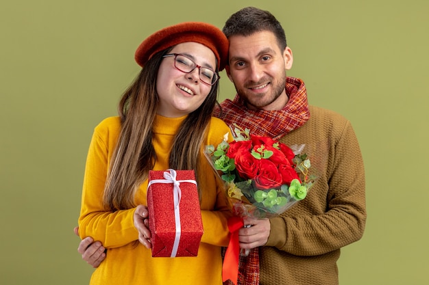 young beautiful couple happy woman in beret with present and man with bouquet of red roses looking at camera smiling happy in love together celebrating valentines day standing over green wall