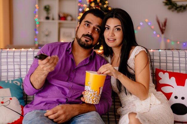 Young and beautiful couple   happy woman and man sitting on a couch with bucket of popcorn watching tv together in decorated room with christmas tree  in the background