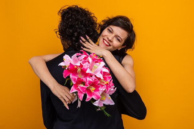 バレンタインを祝う愛で幸せな彼女のボーイフレンドを抱き締めて元気に笑って花の花束を持つ若い美しいカップル幸せな女性