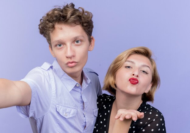 Young beautiful couple hapy in love blowing kisses standing over blue wall