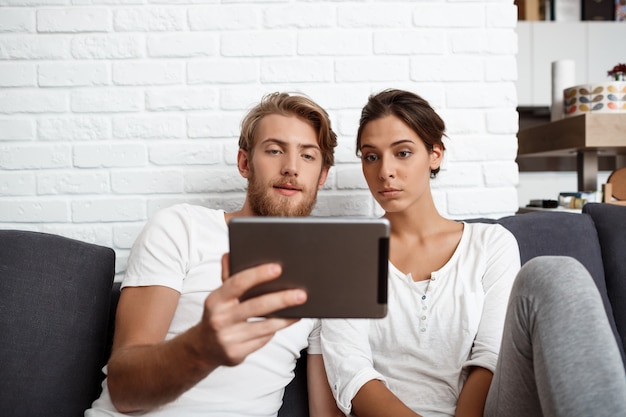 Young beautiful couple looking at tablet sitting on sofa  home.