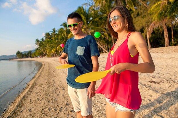Молодая красивая влюбленная пара, играющая в пинг-понг на тропическом пляже, весело, летние каникулы, активная, улыбающаяся, веселая, позитивная