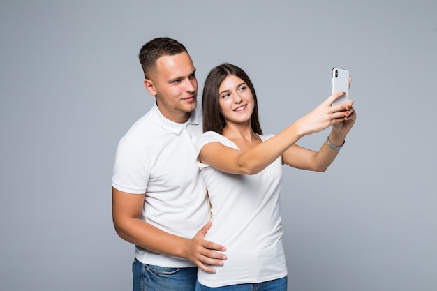 Young beautiful couple in love taking romantic self portrait selfie photo together with mobile phone smiling happy wearing trendy clothes isolated on grey background