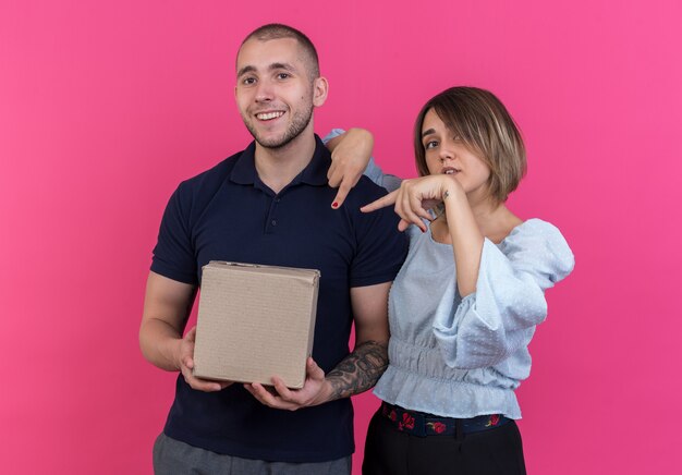 Young beautiful couple man holding cardboard box while his girlfriend pointing with index fingers at box standing