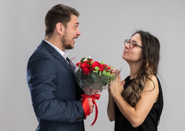 Young beautiful couple man with bouquet of roses looking at his pleased and happy girlfriend happy in love celebrating valentines day over white wall