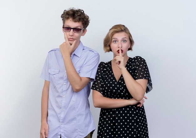 Young beautiful couple man with pensive expression on face and woman making silence gesture with finger on lips standing over white wall