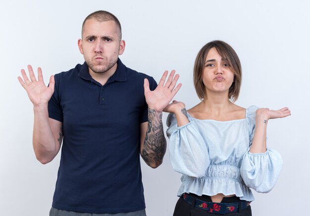 Young beautiful couple man and woman  confused spreading arms to the sides having no answer standing over white wall