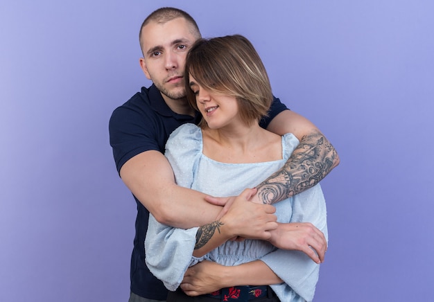 Young beautiful couple man and woman embracing happy in love together smiling standing over blue wall