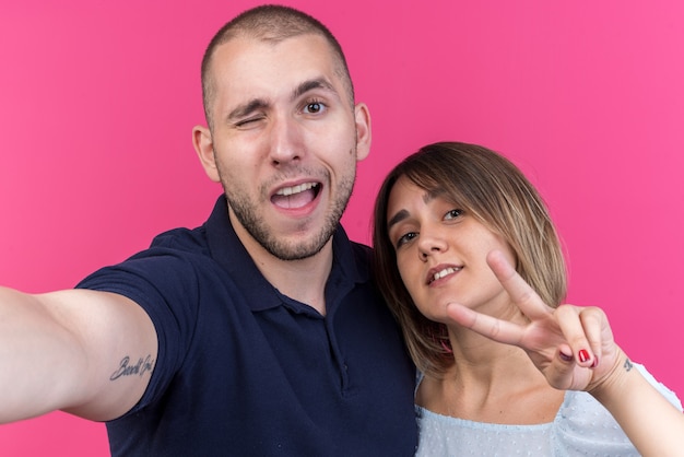 Giovane bella coppia uomo e donna felice e positiva che sorride allegramente mostrando il v-sign in piedi sopra il muro rosa