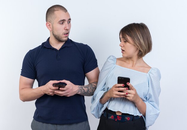 Молодая красивая пара мужчина и женщина, держащая смартфоны, смущают женщину, подозрительно глядя на своего парня, стоящего на смартфоне
