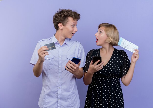 Young beautiful couple man and woman holding smartphones and credit card with air tickets happy and surprised looking at each other over blue wall