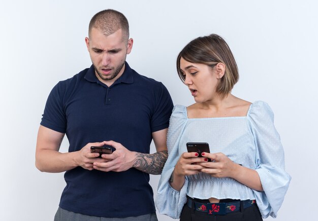 Молодая красивая пара мужчина и женщина, держащая смартфоны, выглядят смущенными и удивленными стоя