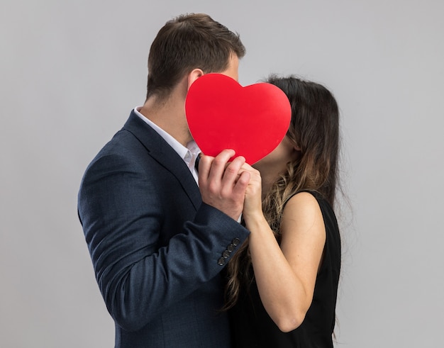 Young beautiful couple man and woman kissing behind red heart happy in love celebrating valentine's day