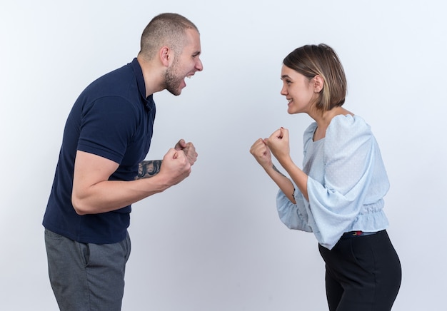 Young beautiful couple man and woman quarreling shouting with clenched fists standing over white wall