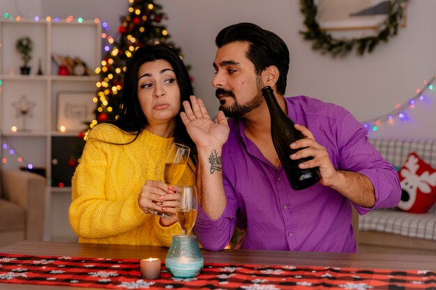 young and beautiful couple man and woman sitting at the table with glasses of champagne happy in love celebrating christmas together in christmas decorated room with christmas tree in the background