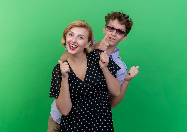 Young beautiful couple man and woman  smiling happy and positive showing thumbs up standing over green wall