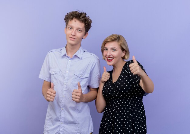 젊은 아름 다운 부부 남자와 여자는 파란색 벽 위에 서 엄지 손가락을 보여주는 미소