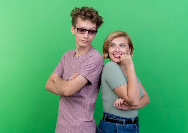 Молодая красивая пара мужчина и женщина, стоящие спиной к спине, счастливые и позитивные, улыбаясь над зеленой стеной