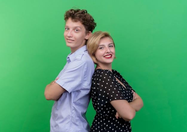 Young beautiful couple man and woman standing back to back  smiling broadly over green wall