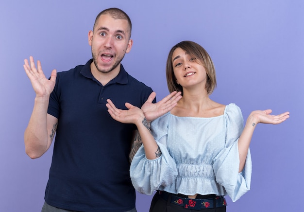 Free photo young beautiful couple man and woman  surprised smiling cheerfully spreading arms to the sides standing over blue wall