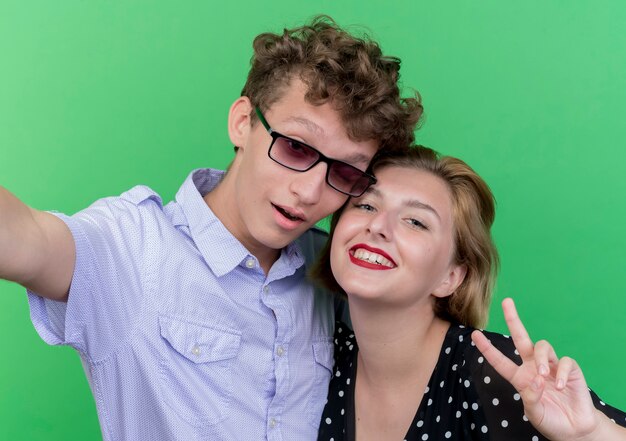 Young beautiful couple man and woman taking selfie  happy and positive showing v-sign standing over green wall