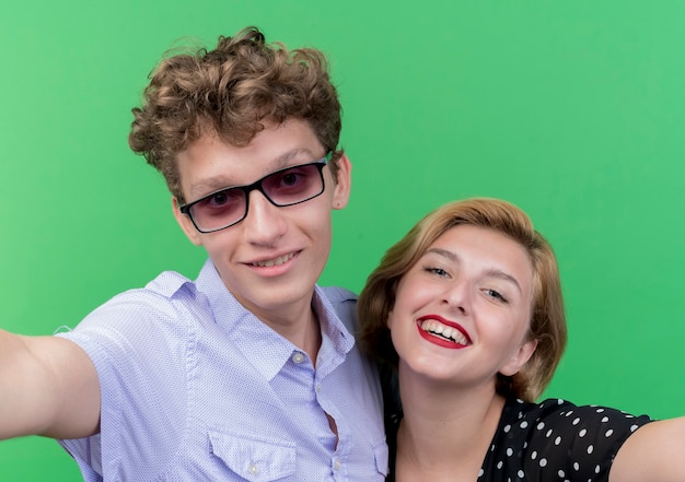 Young beautiful couple man and woman taking selfie happy smiling broadly standing over green wall