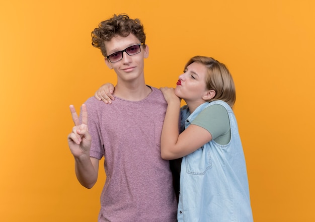 Young beautiful couple man and woman wearing casual clothes happy in love standing together showing v-sign standing over orange wall