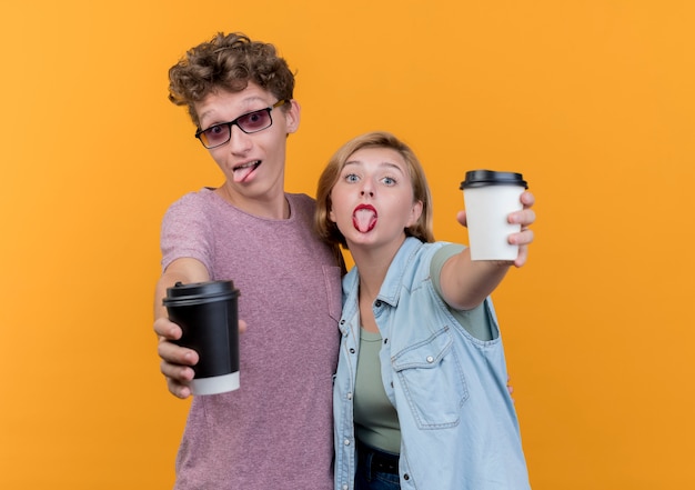 Young beautiful couple man and woman wearing casual clothes showing coffee cups  posing and smiling sticking out tongues standing over orange wall