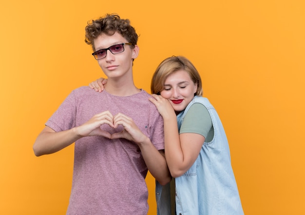 カジュアルな服を着た若い美しいカップルの男性と女性が一緒に立っている男性は、彼のギルフレンドがオレンジ色の壁を越えて彼の肩に頭を傾けている間、心臓のジェスチャーを示しています