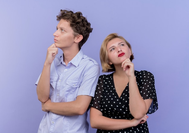 Young beautiful couple man and woman with pensinve expression on faces standing over blue wall