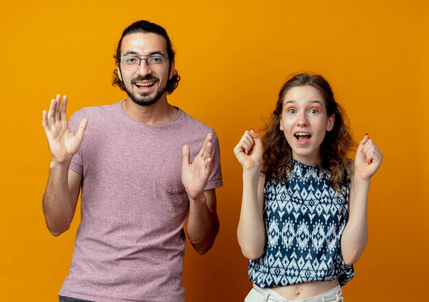 若い美しいカップルの男性と女性はオレンジ色の壁の上に立って腕を上げる幸せで興奮した握りこぶし