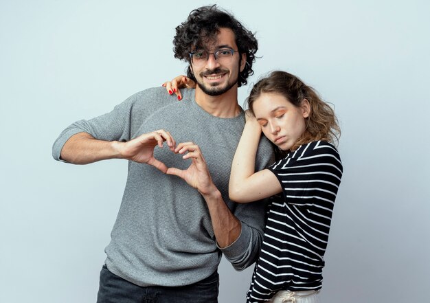 Young beautiful couple man and women happy in love, woman hugging her boyfrind while he making heart gesture with fingers happy and positive over white background
