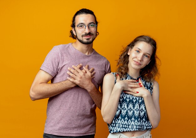 Young beautiful couple man and women looking at camera holding hands on his chests feeling thankful standing over orange background