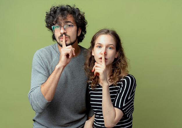 Free photo young beautiful couple man and women looking at camera making silence gesture with fingers on lips over light green background