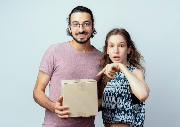 young beautiful couple man and women, man holding box package smiling while his girlfriend pointign with finger at box surprised over white wall