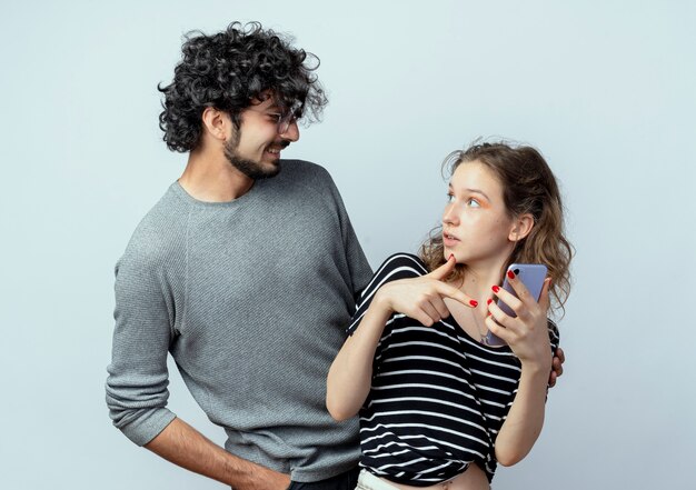 young beautiful couple man and women, man looking at his confused girlfriend which holding smartphone over white wall